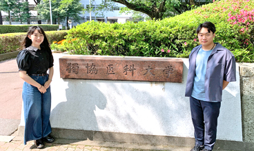 大学の校門の前に立つ増田さんと井上くん
